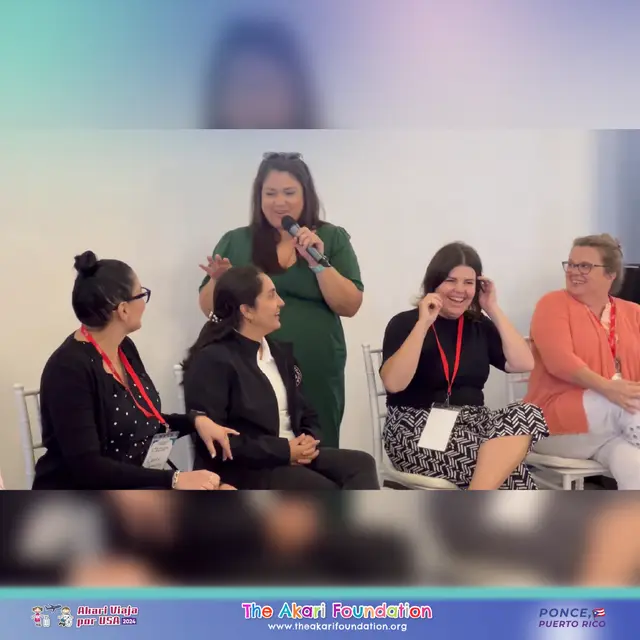 Una mujer de pie habla al micrófono mientras cuatro mujeres sentadas la escuchan y ríen.